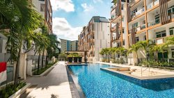 Residential apartments surrounding a blue-tiled swimming pool with palm trees and a clear sunny sky