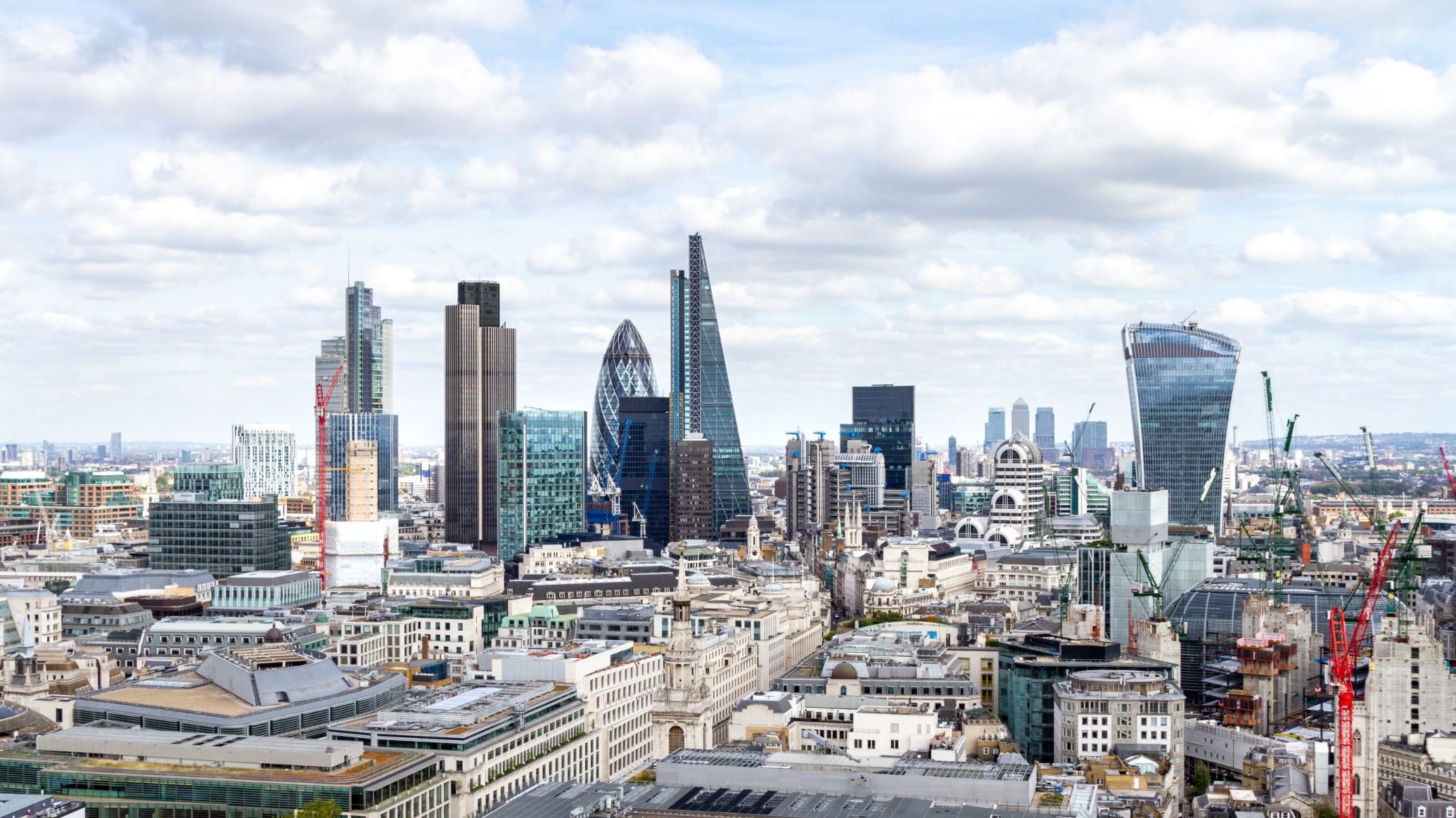 City skyline with tall modern office buildings and construction cranes under a partly cloudy sky