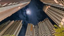 High-rise residential blocks with lit windows under a cloudy night sky and a bright moon