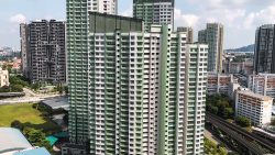 High-rise public housing blocks with green and white façades near a train track and open field under a partly cloudy sky
