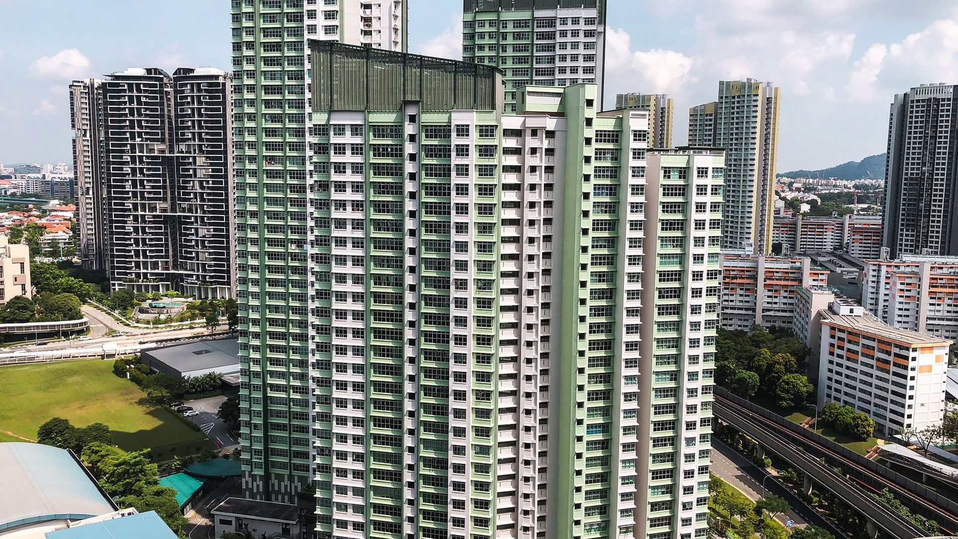 High-rise public housing blocks with green and white façades near a train track and open field under a partly cloudy sky