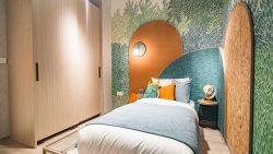 Single bed with colourful cushions against a textured headboard and leafy wallpaper in Mori bedroom