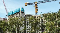 Unfinished concrete residential building with blue safety nets and tower cranes surrounded by trees under clear sky