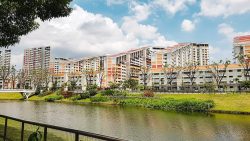 Housing blocks beside a river with a landscaped park and trees under a partly cloudy sky