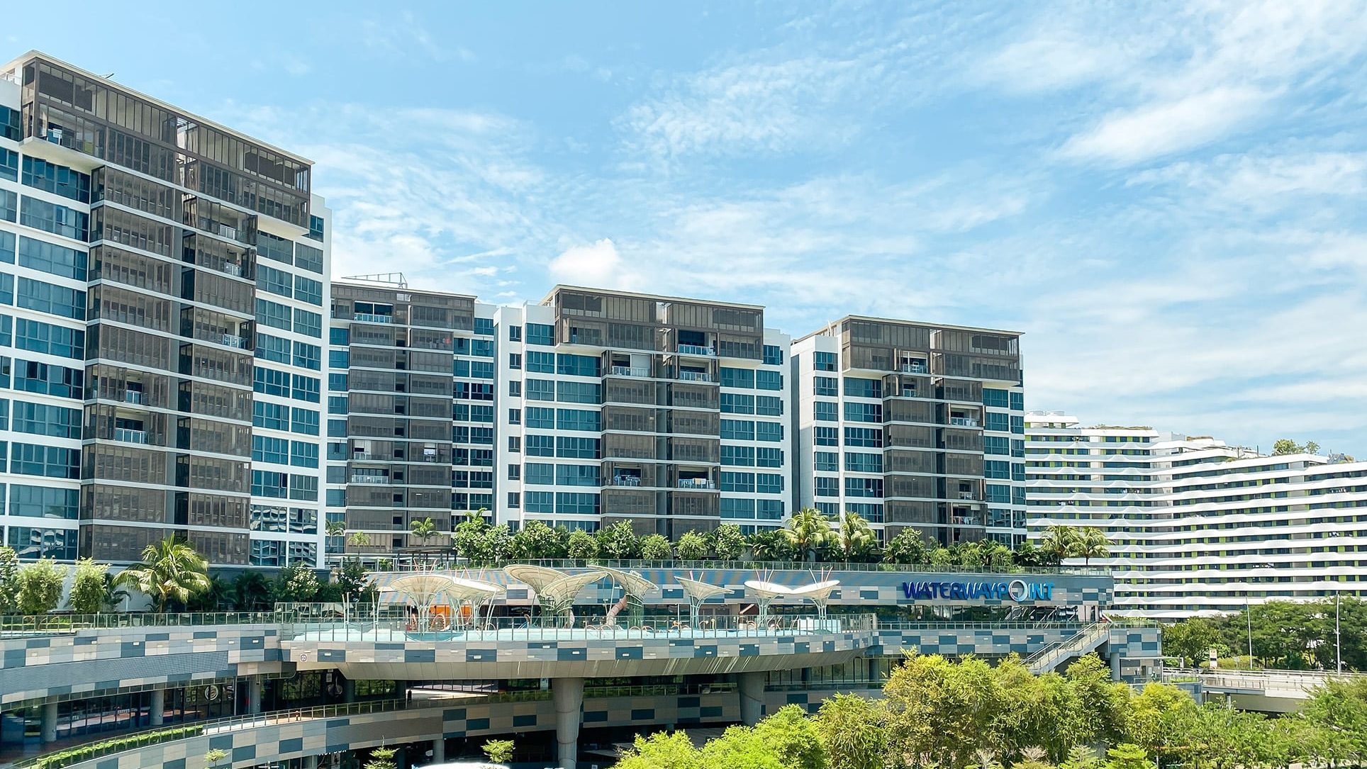 Watertown apartment blocks with blue-tinted windows behind Waterway Point mall and greenery under a blue sky