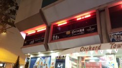 Night view of a retail shopfront with illuminated signage and a partial Orchard Town Centre sign above the entrance