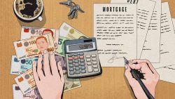 Hands signing mortgage documents on wood table with calculator, keys, currency notes and coffee cup
