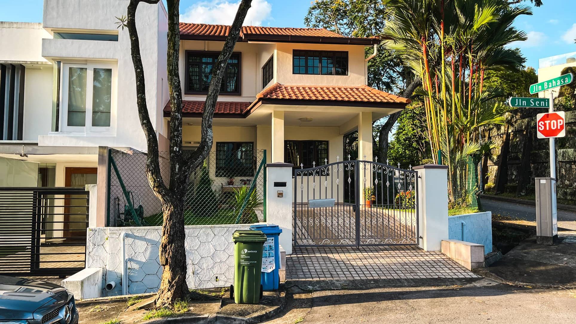 Two-storey terrace house with a gated driveway and recycling bins near street signs and trees