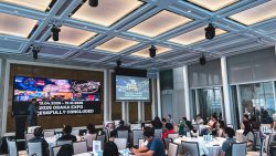 Conference room with attendees seated at round tables and screens displaying 2025 Osaka Expo information