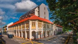 Heritage two-storey shophouses with yellow windows and closed shutters on a sunlit street corner with parked cars and trees