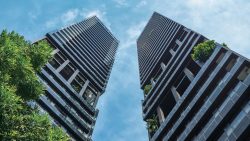 Two tall residential towers with glass balconies and greenery on upper floors against blue sky