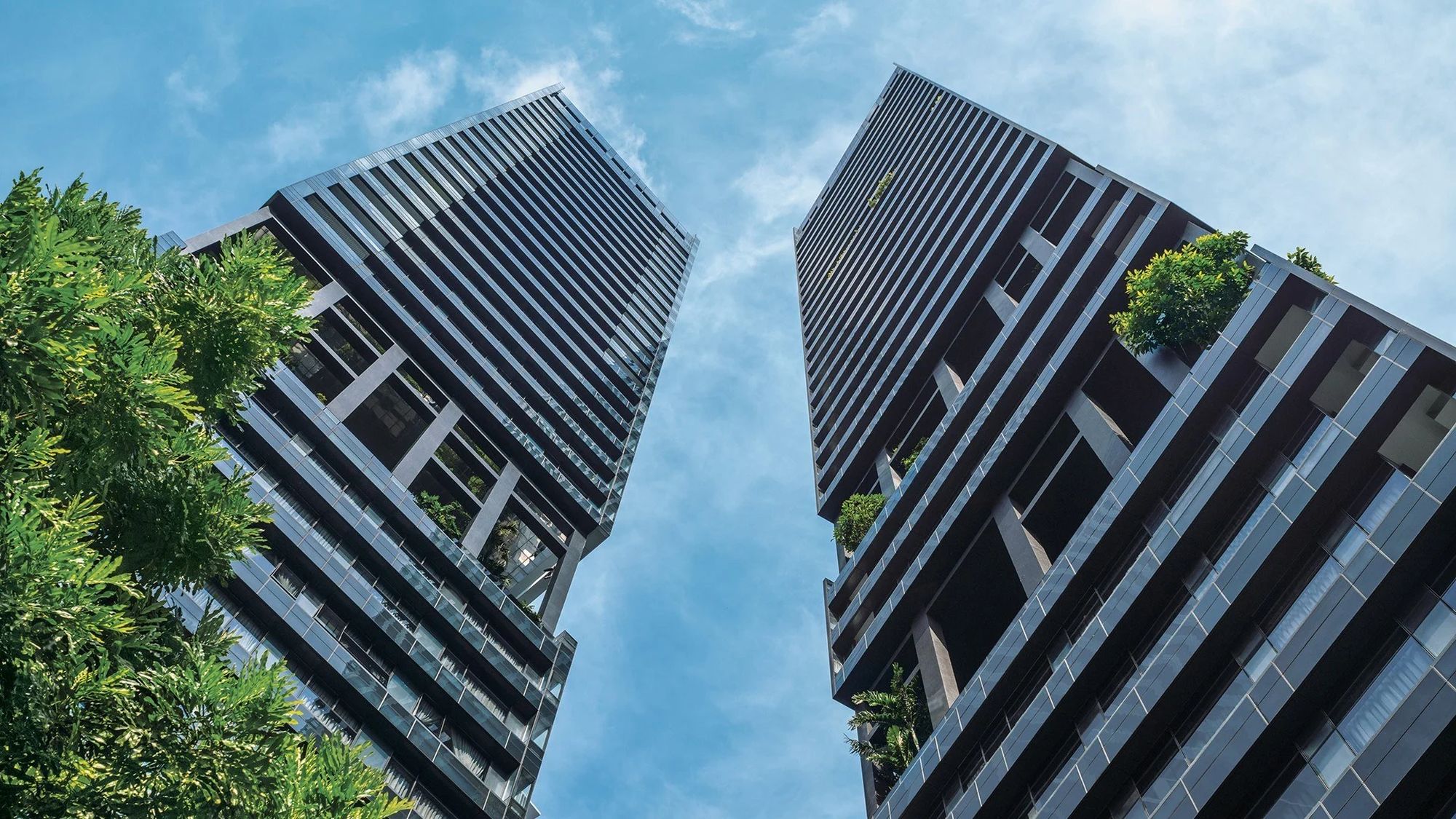 Two tall residential towers with glass balconies and greenery on upper floors against blue sky