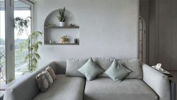 Living room with grey sofa, pastel green cushions, plush toys, built-in wall shelf, and tall indoor plant near window