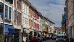 Shophouses with colourful shutters and tiled roofs line a street with cars parked along the roadside